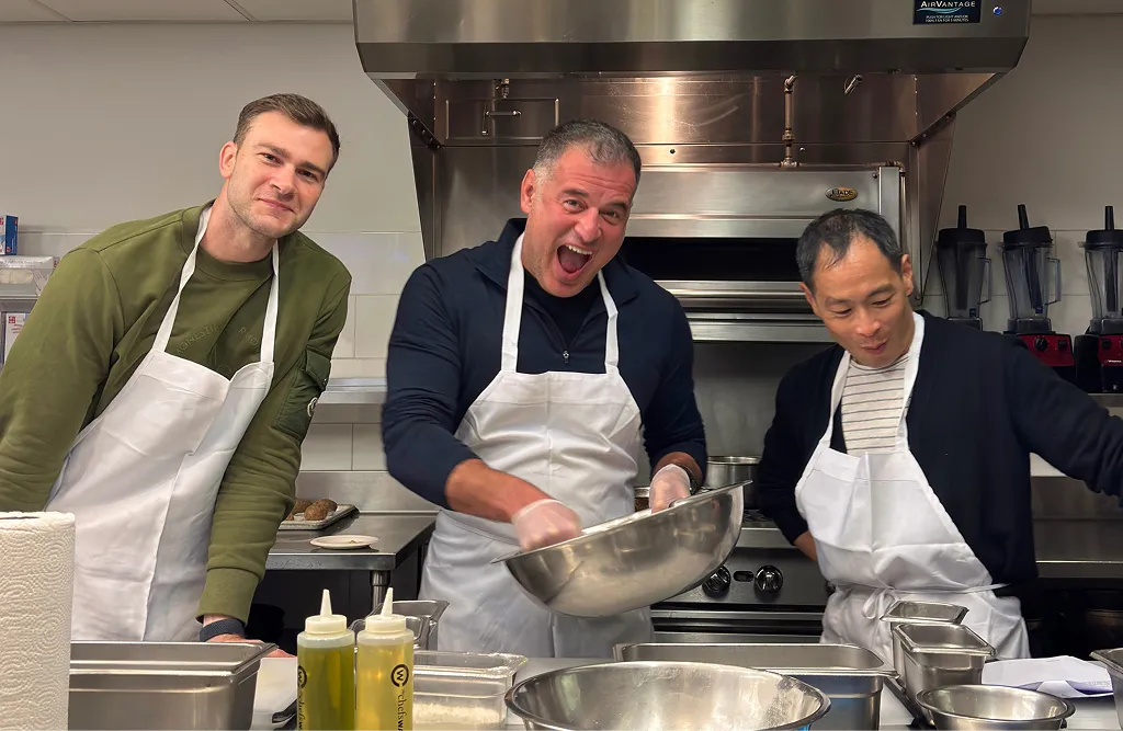 Three men in aprons cooking together in a commercial kitchen, one stirring a large metal bowl.