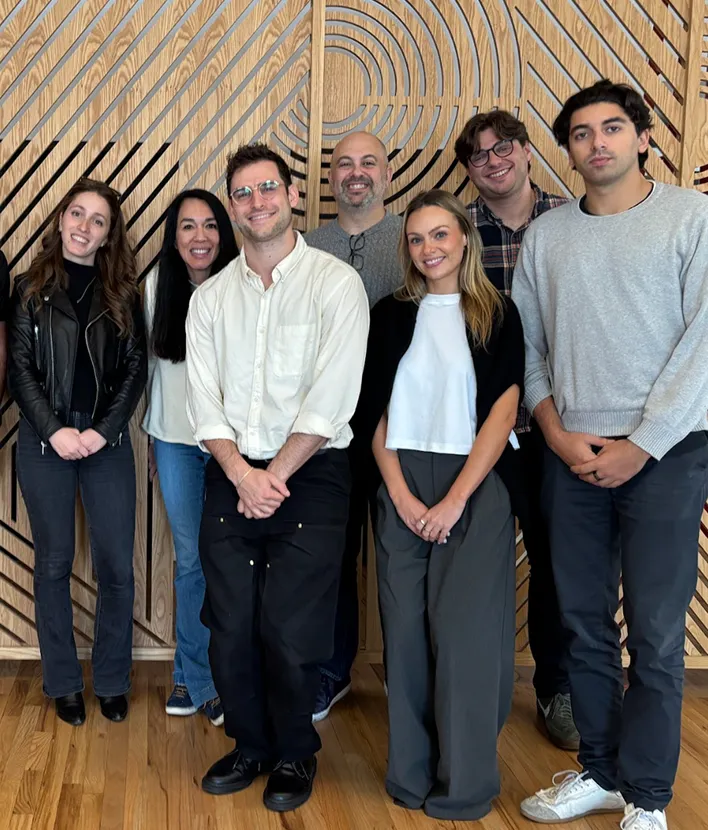 Group of seven diverse people smiling and standing in front of a wooden wall with geometric pattern.