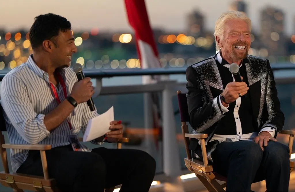 Two men seated outdoors in evening, smiling and holding microphones during a discussion or interview.