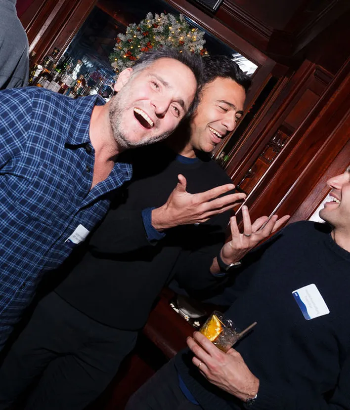 Three men laughing and chatting inside a wood-paneled room with a decorated Christmas wreath in the background.
