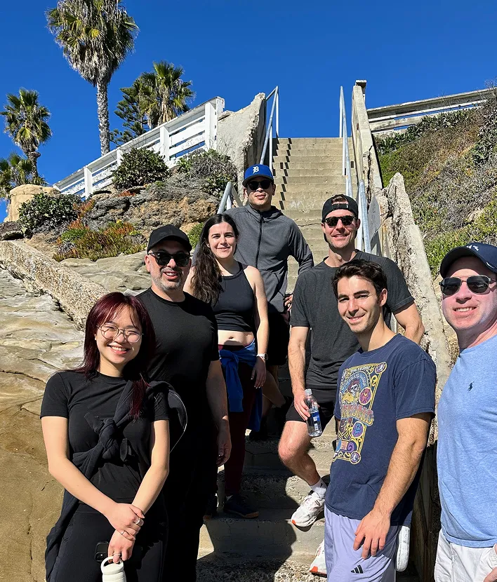 Group of seven people smiling on outdoor stairs with blue sky and palm trees in the background.