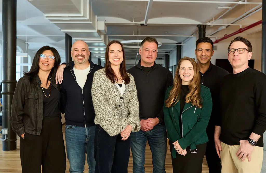 Seven diverse adults standing together inside a modern office space, smiling at the camera.